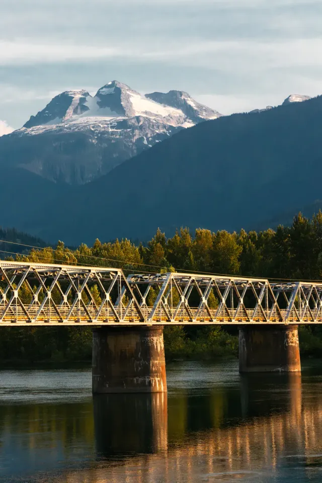 A steel truss bridge crossing a river below forested mountain slopes in Alberta