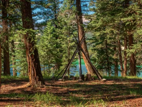 A traditional teepee frame standing among tall pines beside a lake